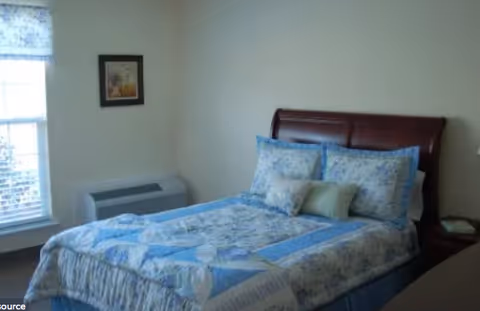 A bedroom with a bed made up with blue and white floral bedding, a wooden headboard, a window with a floral valance, a framed picture on the wall, and a small air conditioning unit below the window.