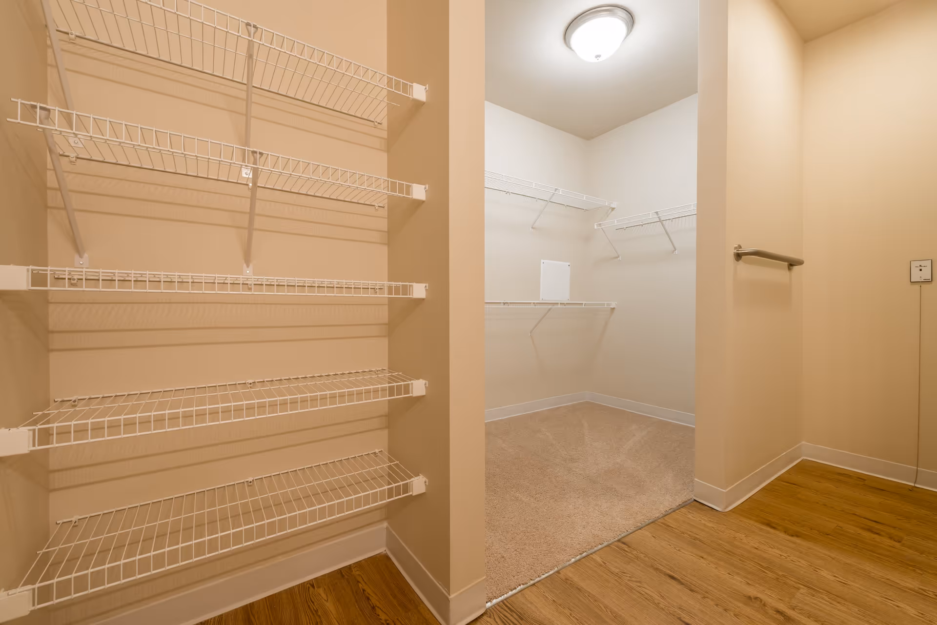 Empty walk-in closet with beige walls, white wire shelving on the walls, a ceiling light fixture, and wood flooring transitioning to carpet inside the closet area.