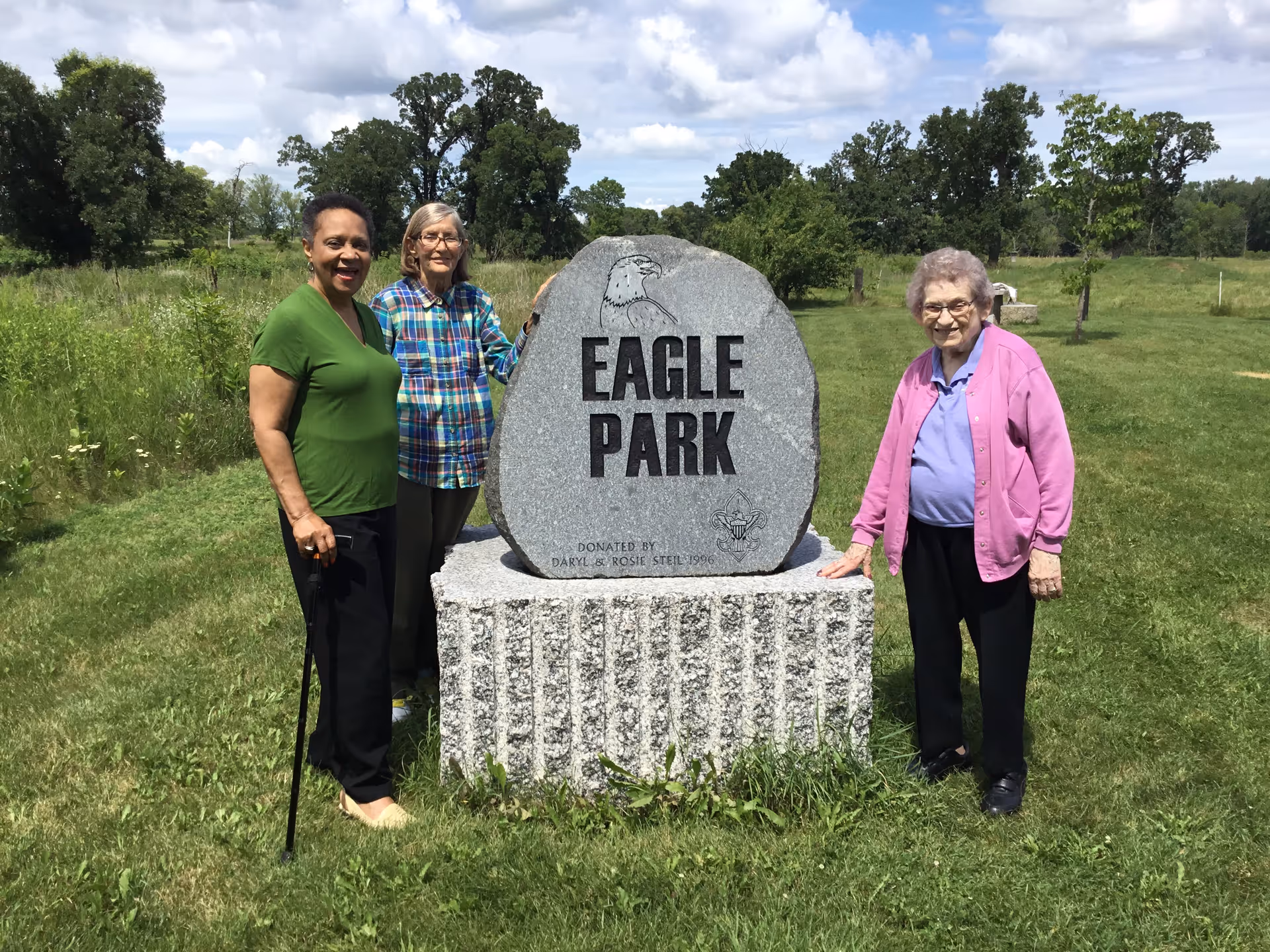 Three elderly women standing outdoors on grass around a large stone monument that reads 'Eagle Park'. The sky is partly cloudy and there are trees and greenery in the background.