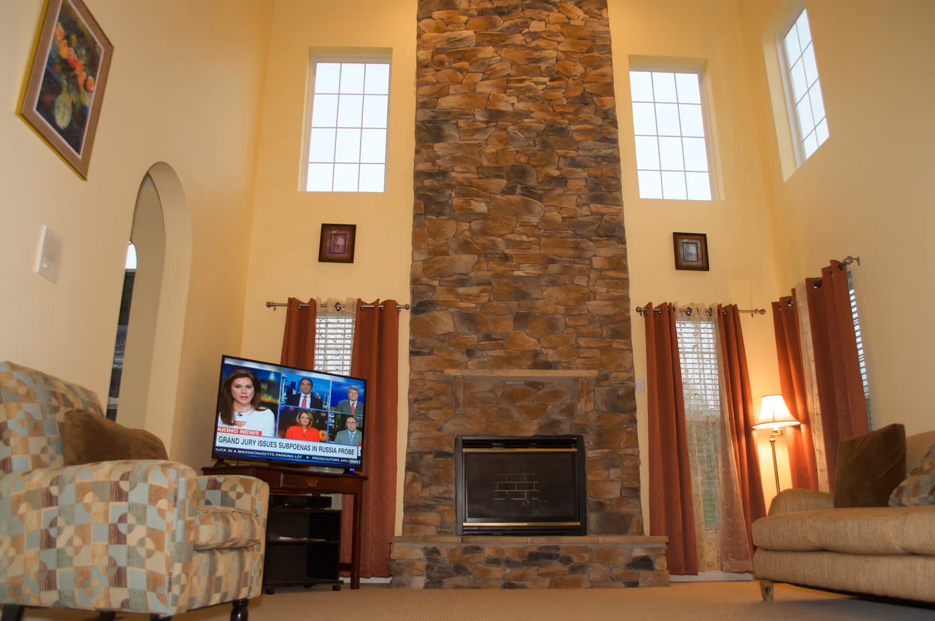 A cozy living room with a tall stone fireplace centered between two sets of windows with red curtains. There is a patterned armchair on the left and a beige sofa with brown cushions on the right. A TV on a stand is positioned to the left of the fireplace, displaying a news broadcast. A floor lamp is lit near the sofa on the right side. The walls are painted a light yellow color and there is a framed painting on the left wall.