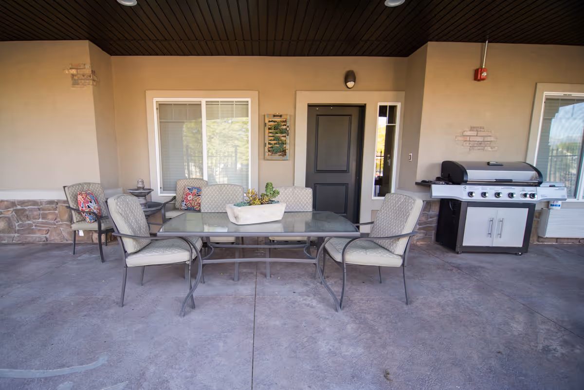 Outdoor patio area with a glass-top table surrounded by four cushioned chairs. There is a rectangular planter with succulents on the table. Behind the table, there is a black door, two windows with white frames, a wall-mounted light fixture, and a large stainless steel grill on the right side. The patio floor is concrete, and the walls are beige with some stone accents.