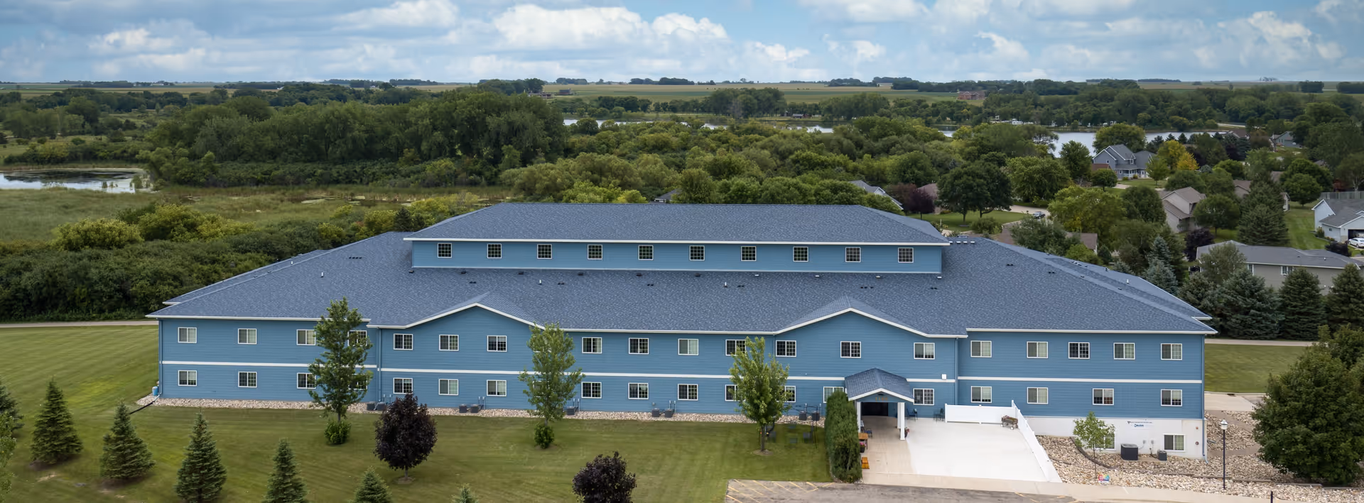 A large, blue, two-story building with multiple windows surrounded by green trees and grass under a partly cloudy sky, viewed from the back or side with a paved area and entrance visible.