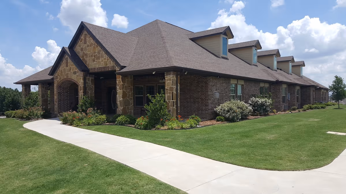 Single-story brick and stone building with a sloped roof, a curved concrete walkway, and manicured lawn and shrubs under a partly cloudy sky.