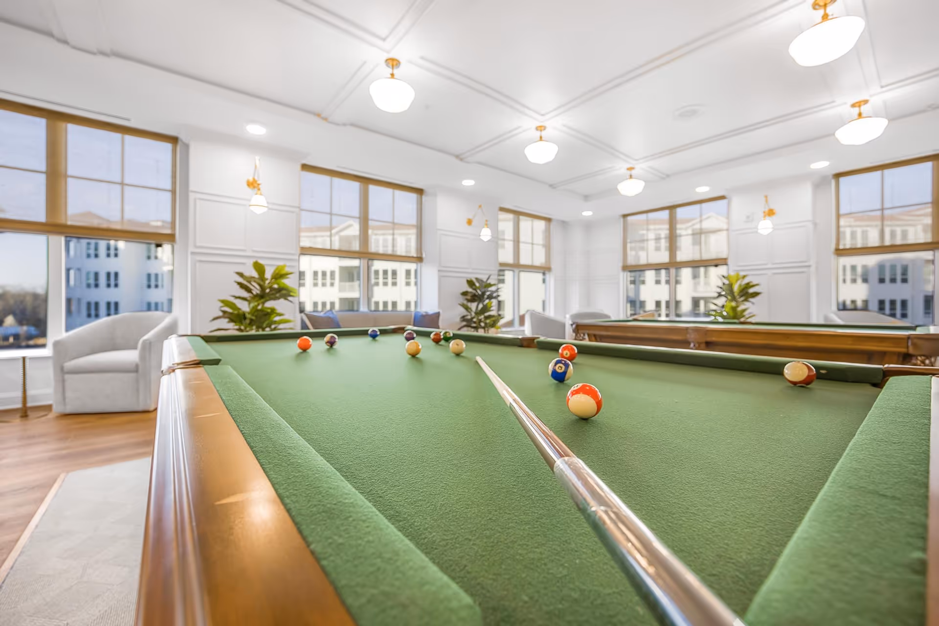 A bright and spacious room with large windows, featuring a green felt pool table with billiard balls and a cue stick positioned for a shot. The room has white walls with decorative molding, wooden flooring, several potted plants, and comfortable seating areas with armchairs.