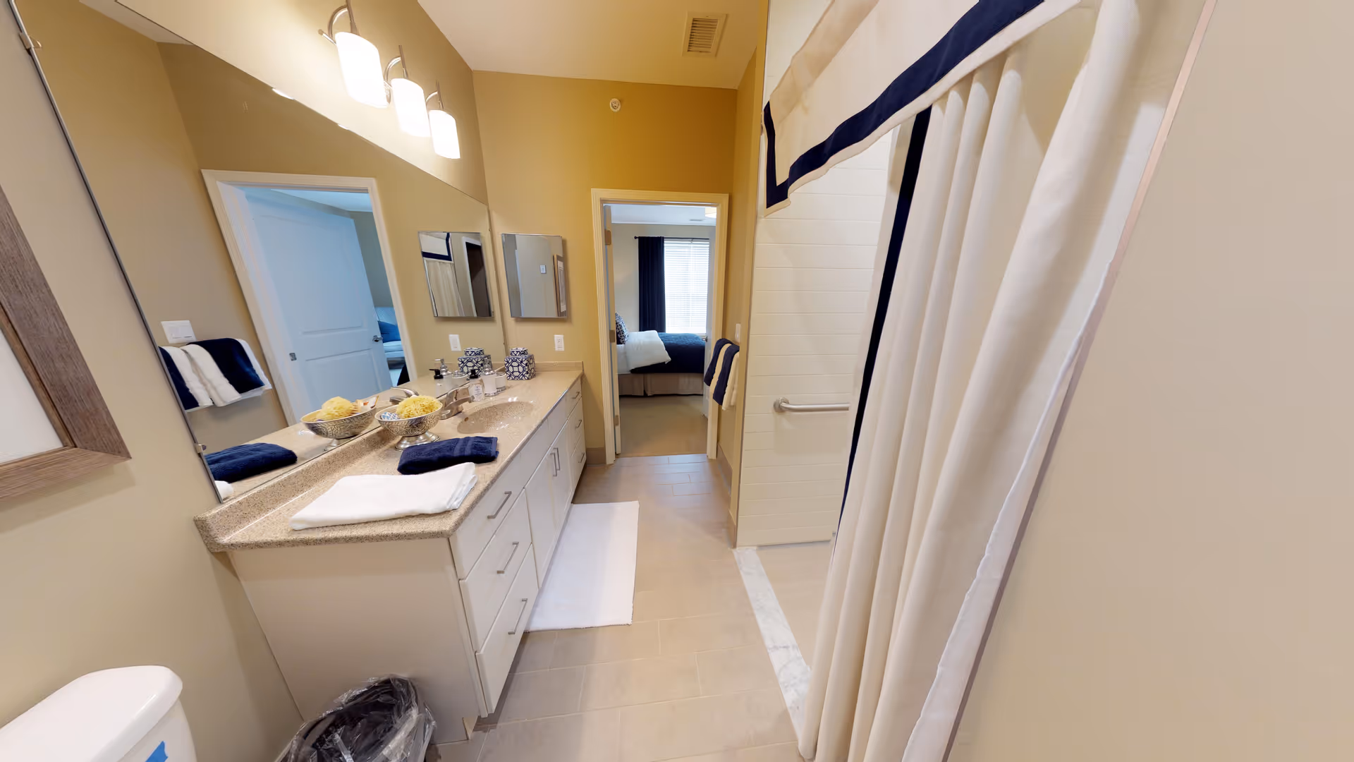 A clean and well-lit bathroom featuring a long countertop with two sinks, a large mirror above, and neatly folded towels. A shower with a white curtain is visible on the right side. The bathroom opens into a bedroom with a bed and window with curtains.