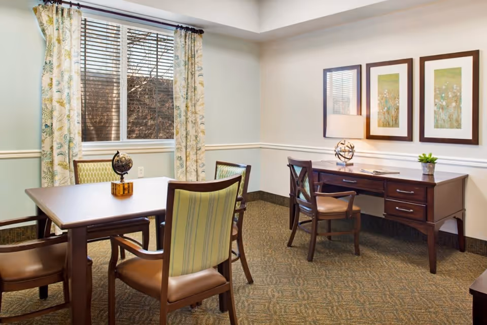 A well-lit room with a wooden table surrounded by four chairs with green and beige striped upholstery. A small globe sits on the table. Against the wall is a wooden desk with a matching chair, a decorative lamp, a small plant, and two framed floral artworks hanging above. A window with floral curtains lets in natural light.