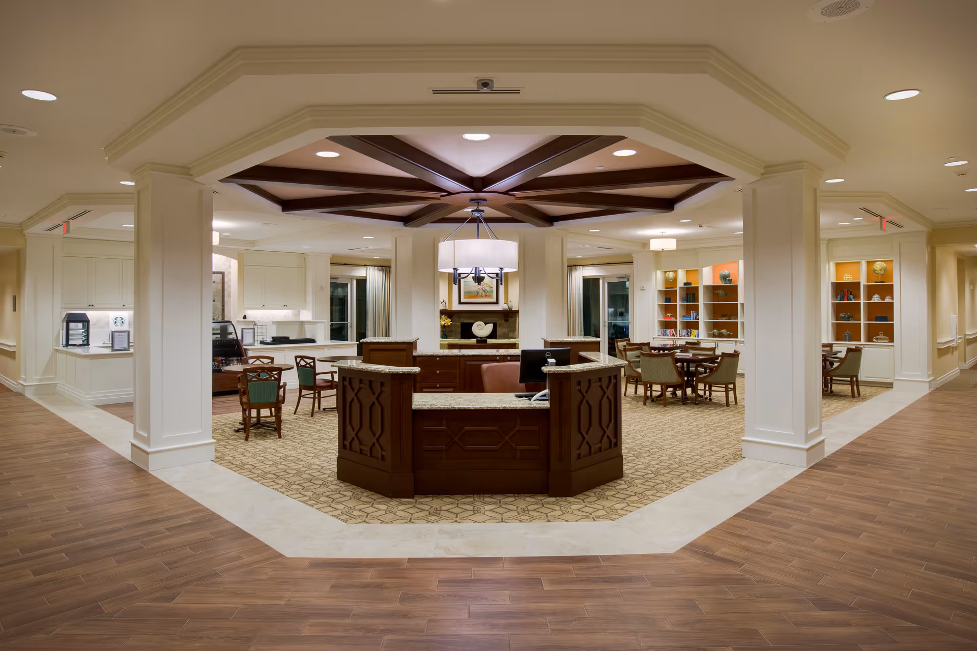 Interior view of a senior living facility reception area with a central wooden desk, surrounded by seating areas with tables and chairs. The ceiling features a decorative wooden beam design with a hanging light fixture. The space is well-lit with recessed lighting and has a combination of wood and carpet flooring. Shelves with decorative items and books are visible in the background.