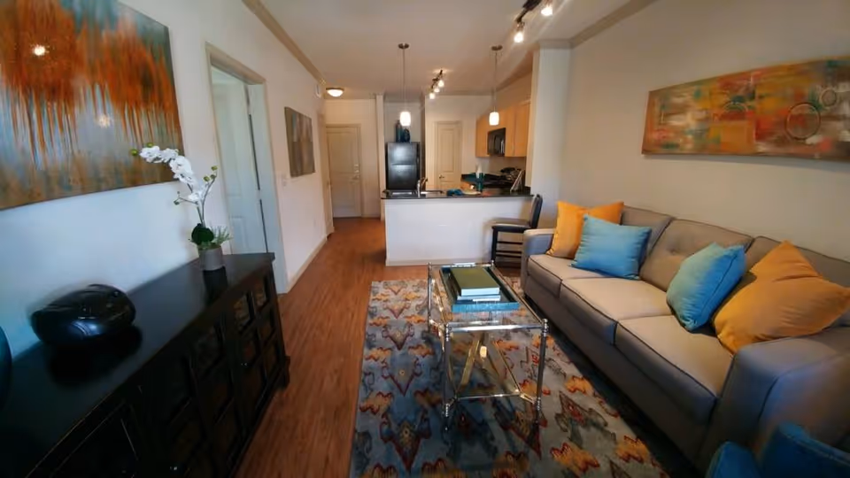 A cozy living room area with a gray sofa adorned with blue and orange pillows, a glass coffee table with books on top, a colorful patterned rug, and a black sideboard with a white orchid plant and a black decorative item. The room opens into a kitchen area with wooden cabinets, a black refrigerator, and a breakfast bar with two chairs. The walls are decorated with abstract paintings, and the floor is wooden.