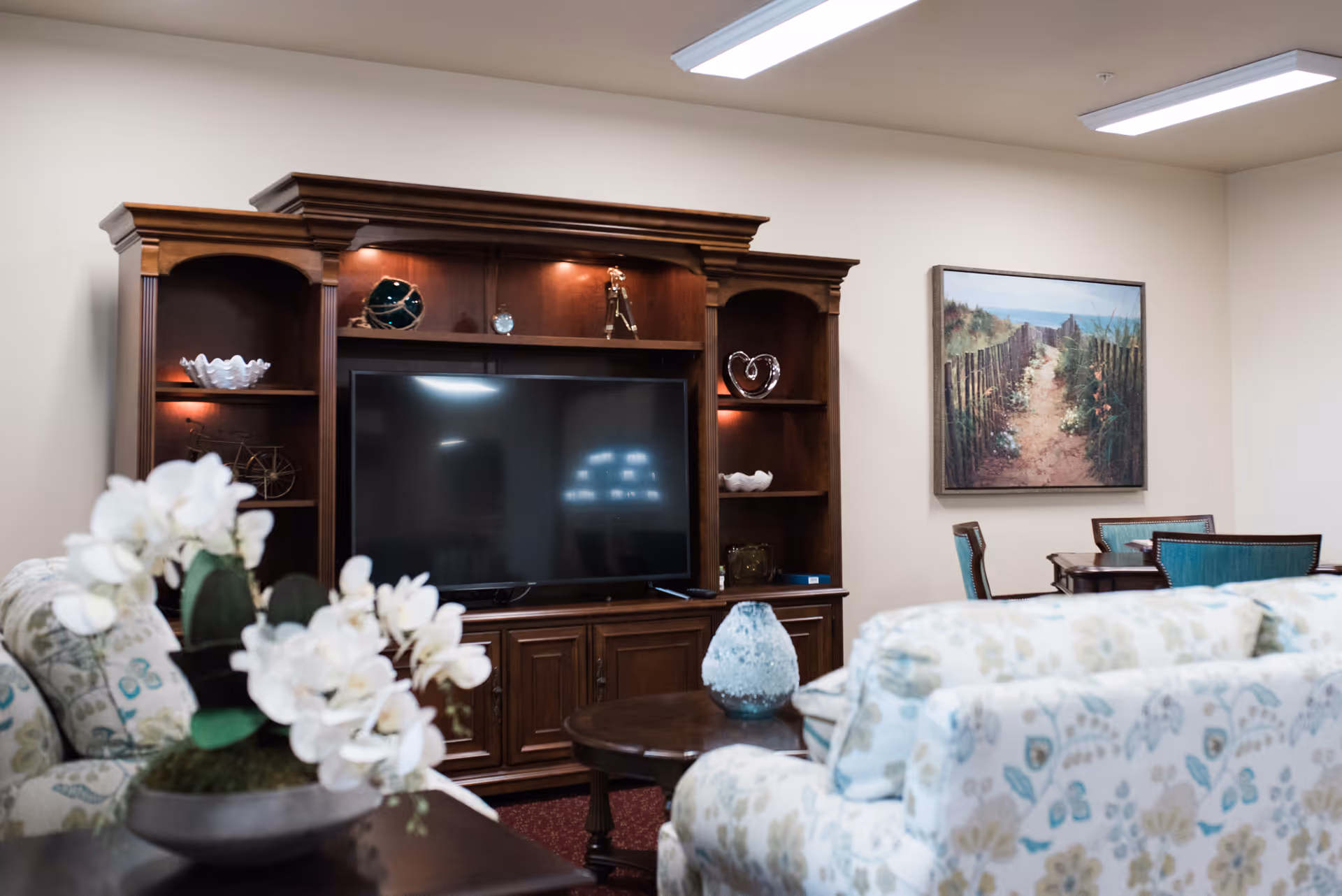 A cozy living room area with floral patterned sofas facing a large wooden entertainment center holding a flat-screen TV. The entertainment center has shelves with decorative items and is illuminated by small lights. A round wooden coffee table with a decorative vase is in front of the sofas. On the wall, there is a framed painting of a sandy path leading through a fence towards the ocean. The room has soft lighting and a calm, inviting atmosphere.