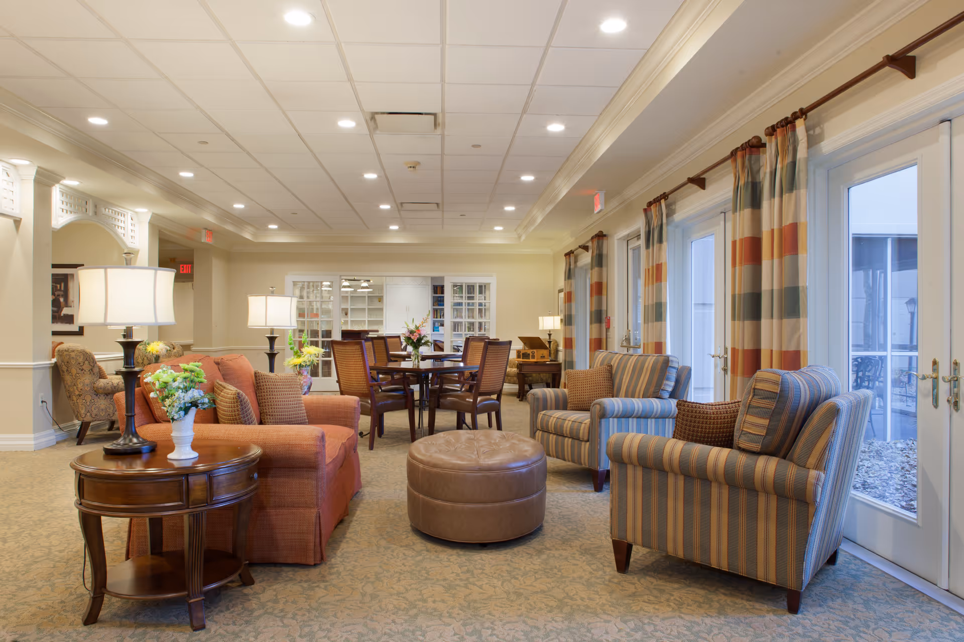 Sunlit senior living common lounge with upholstered sofas and armchairs, tables, lamps, and a dining area by glass doors.
