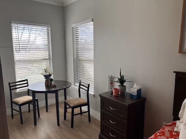 A small dining area with a round table and two chairs next to two windows with blinds. A dark wooden dresser with three drawers is beside the table, holding a small plant, a tissue box, a candle, and a decorative sign. Part of a bed with a red and white blanket is visible on the right side.