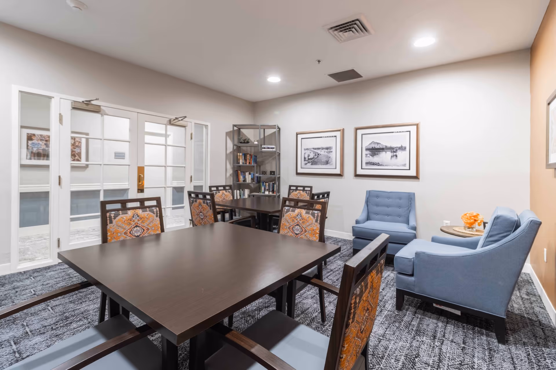 A bright communal room with dark wood tables and patterned chairs, two blue armchairs, framed artwork, and a bookshelf.