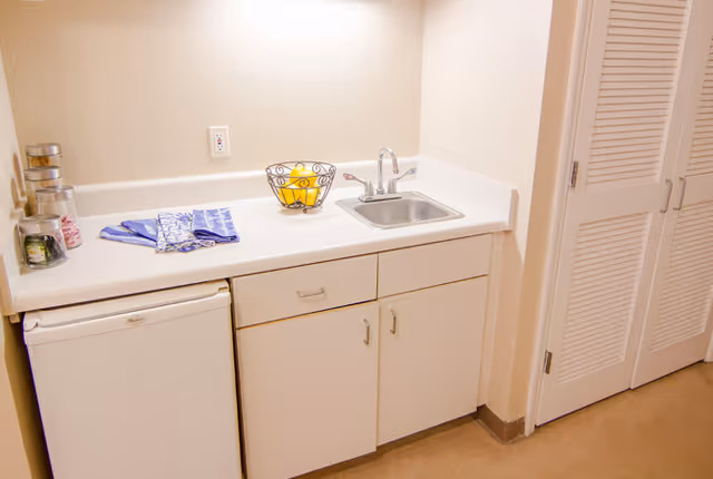 A small kitchenette area with a white countertop, a stainless steel sink with a faucet, a mini refrigerator, and white cabinets below. On the countertop, there are three glass jars, two folded blue and white dish towels, and a wire fruit basket containing lemons. To the right, there is a set of white louvered closet doors.