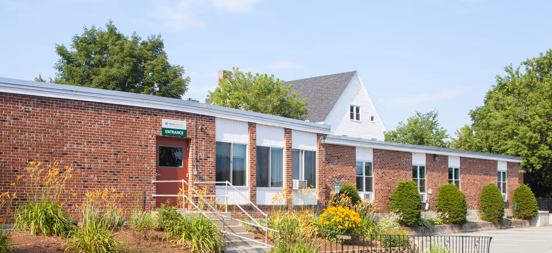 Single-story brick senior living building with an entrance, windows and landscaped flowerbeds under a clear sky.