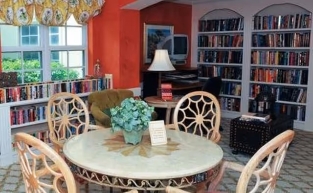 A cozy library or reading room with a round table and four decorative chairs in the foreground. A potted plant and a small sign are on the table. In the background, there are bookshelves filled with books, a green armchair, a small table with a lamp, and a TV on a stand. The room has a window with patterned curtains and a carpeted floor.