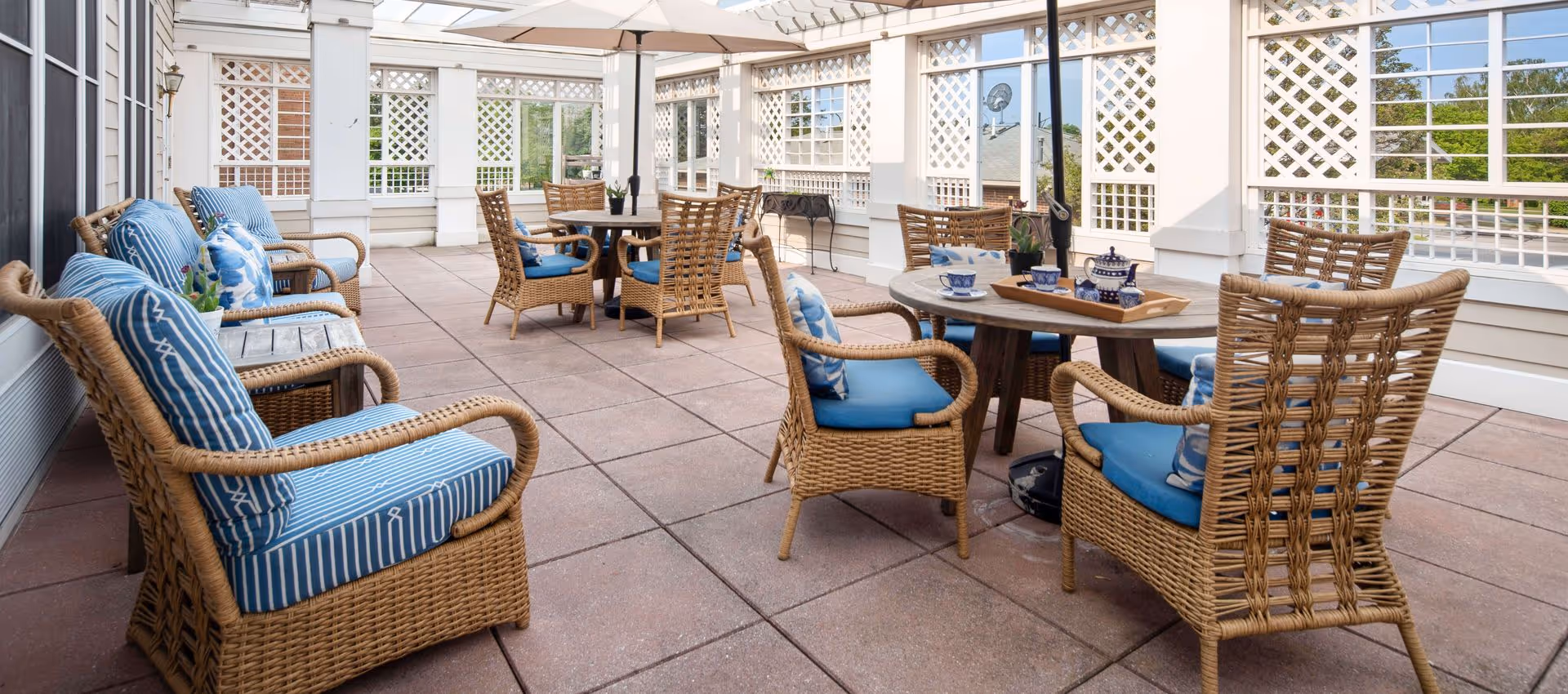 Outdoor patio area with wicker chairs and tables, featuring blue cushions and pillows. The space is enclosed with white lattice panels and has a tiled floor, creating a bright and inviting seating area.