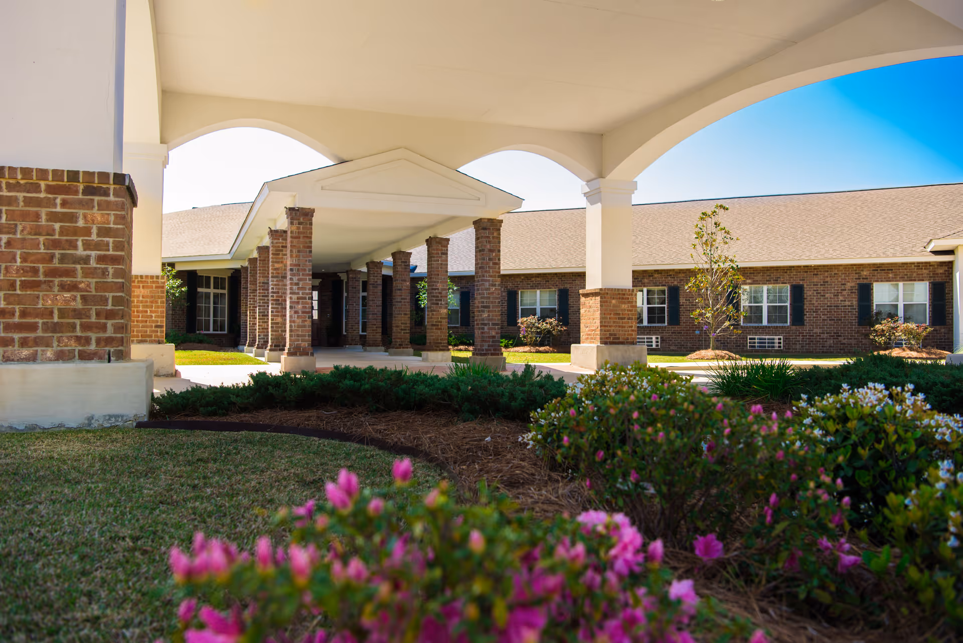 View of the exterior entrance area of a brick building with a covered walkway supported by brick columns. There are landscaped bushes and flowers in the foreground under a clear blue sky.