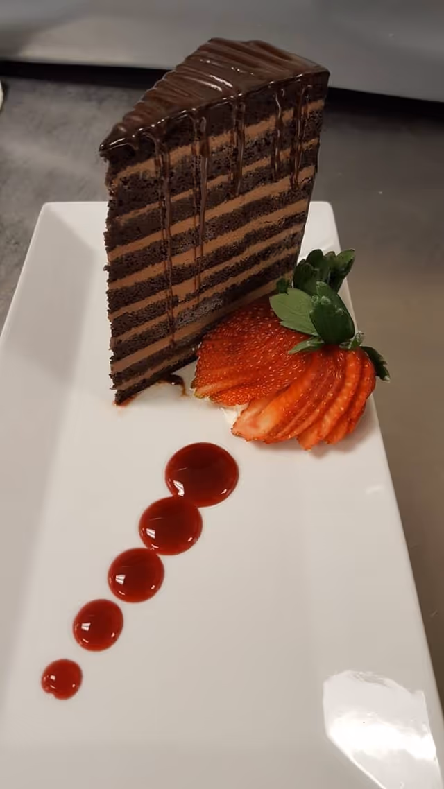 A tall slice of layered chocolate cake with chocolate drizzle on top, served on a white rectangular plate with thinly sliced strawberries and a line of red sauce dots.