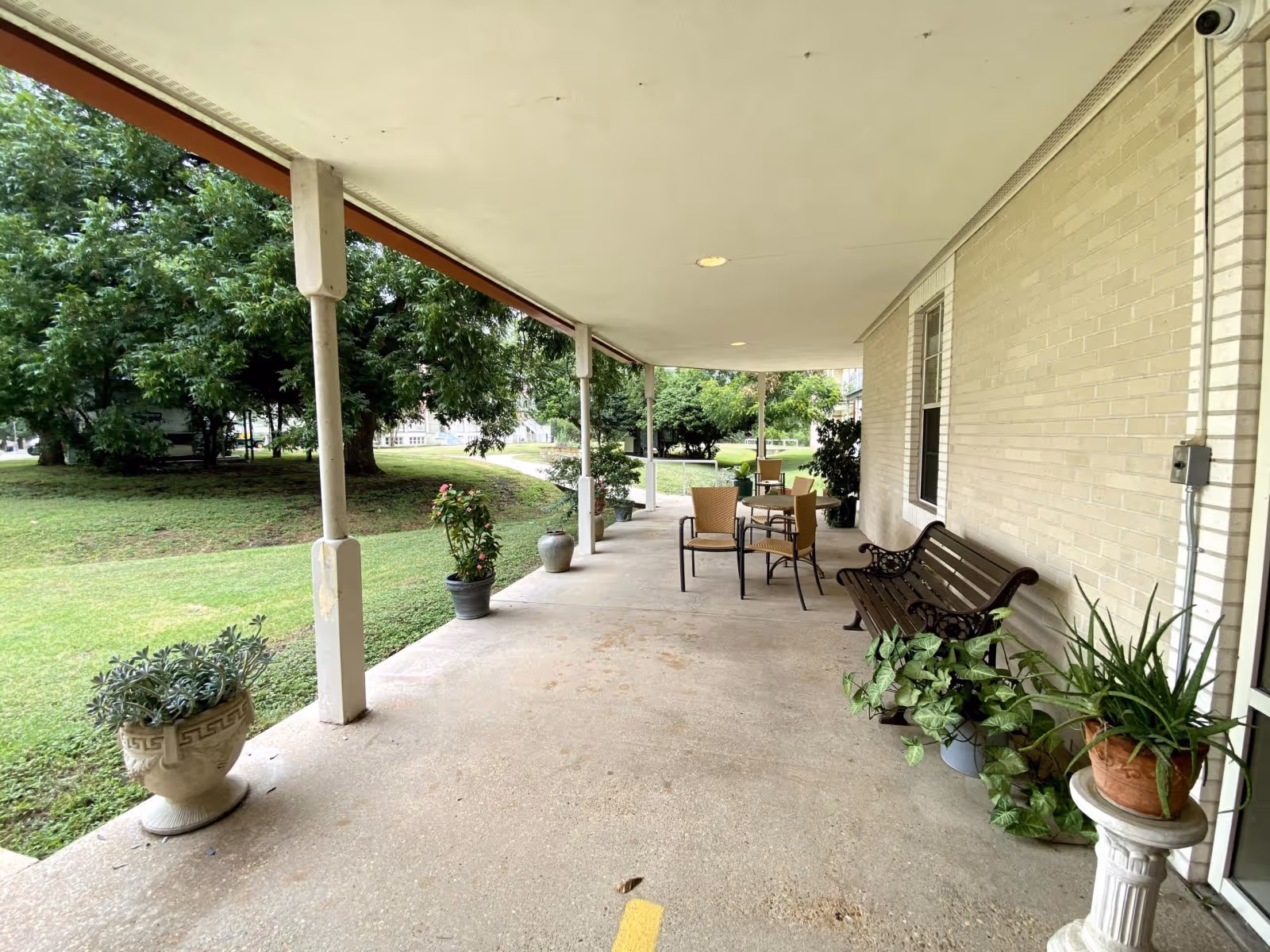 Covered porch with a bench, chairs and potted plants overlooking a grassy yard and trees.