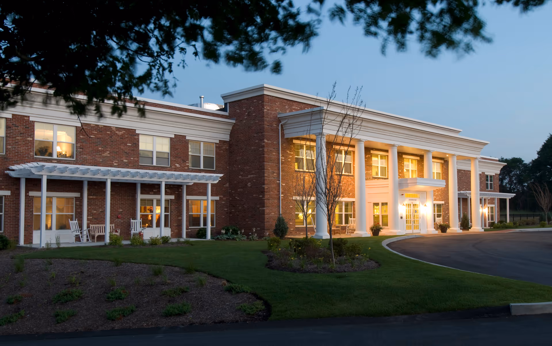 Exterior view of a two-story brick assisted living facility with white columns and a covered entrance, surrounded by a well-maintained lawn and small trees during dusk.
