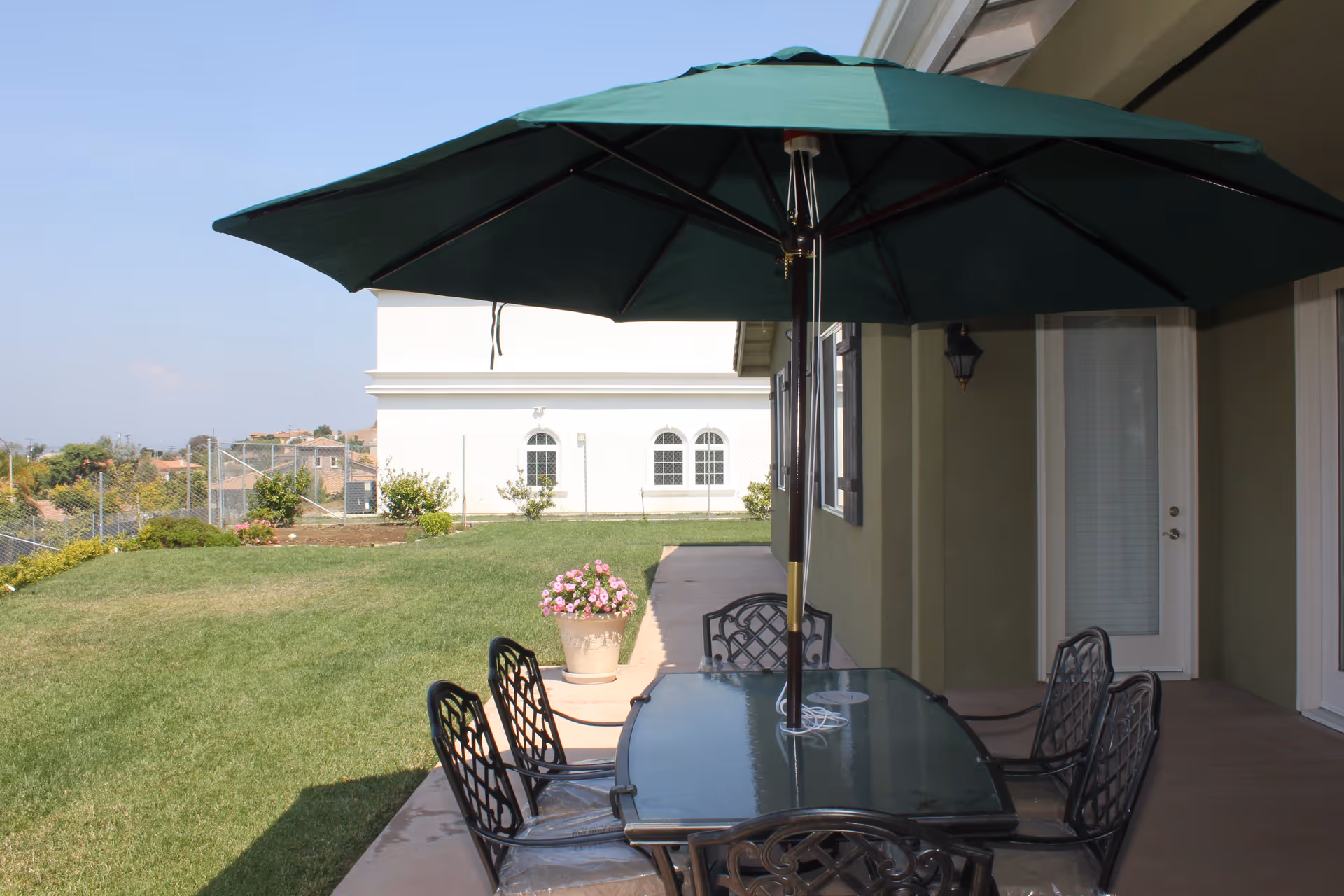Outdoor patio with a glass-top table and metal chairs under a large green umbrella next to a grassy yard and building.