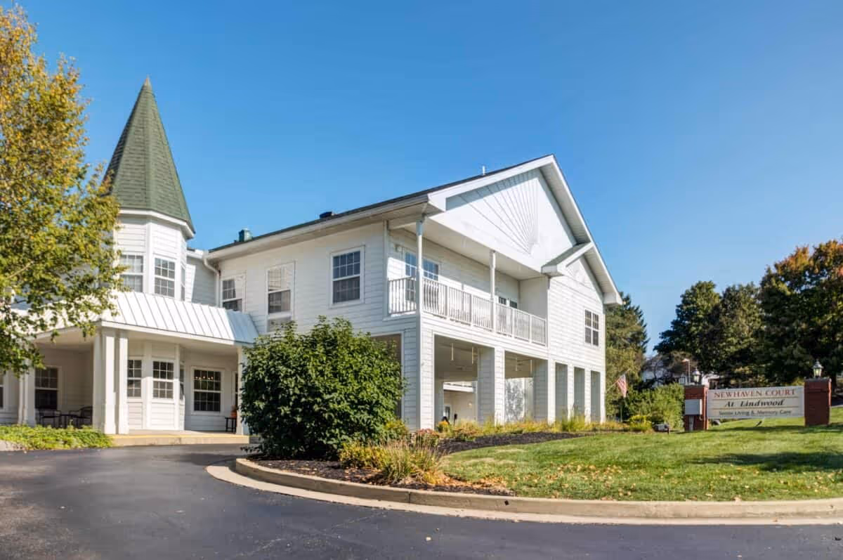 Exterior view of a two-story white building with a pointed turret, surrounded by greenery and a clear blue sky. There is a curved driveway leading to the entrance and a sign on the lawn that reads 'Newhaven Court At Lindwood Senior Living & Memory Care.'