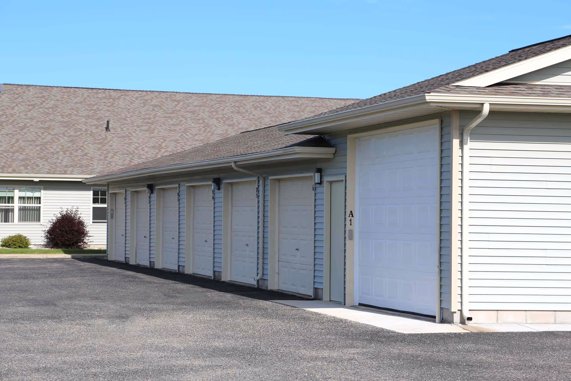 Exterior view of a row of garage doors attached to a light gray building with a shingled roof under a clear blue sky.