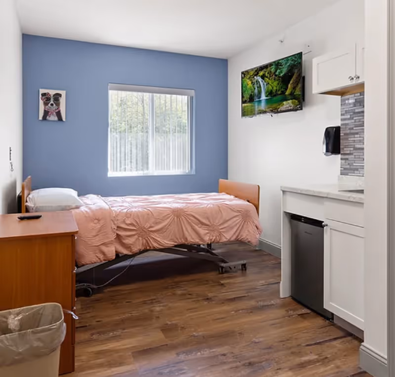 A small, clean bedroom with a single bed covered in a pink comforter, a wooden nightstand with a remote control, a window with vertical blinds, a wall-mounted TV displaying a waterfall scene, and a kitchenette area with white cabinets and a small refrigerator.