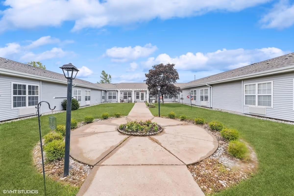 Outdoor courtyard area of a senior living facility with a circular concrete patio featuring a small garden in the center, surrounded by green grass and shrubs. The courtyard is flanked by single-story buildings with gray siding and white-framed windows under a blue sky with scattered clouds.