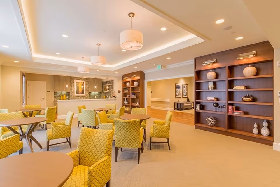 A well-lit common area with multiple round tables surrounded by yellow patterned chairs. The room features a built-in wooden bookshelf with decorative items, a small counter area with a stone backsplash, and modern ceiling lights. The flooring is a mix of carpet and wood, and the walls are painted in neutral tones.