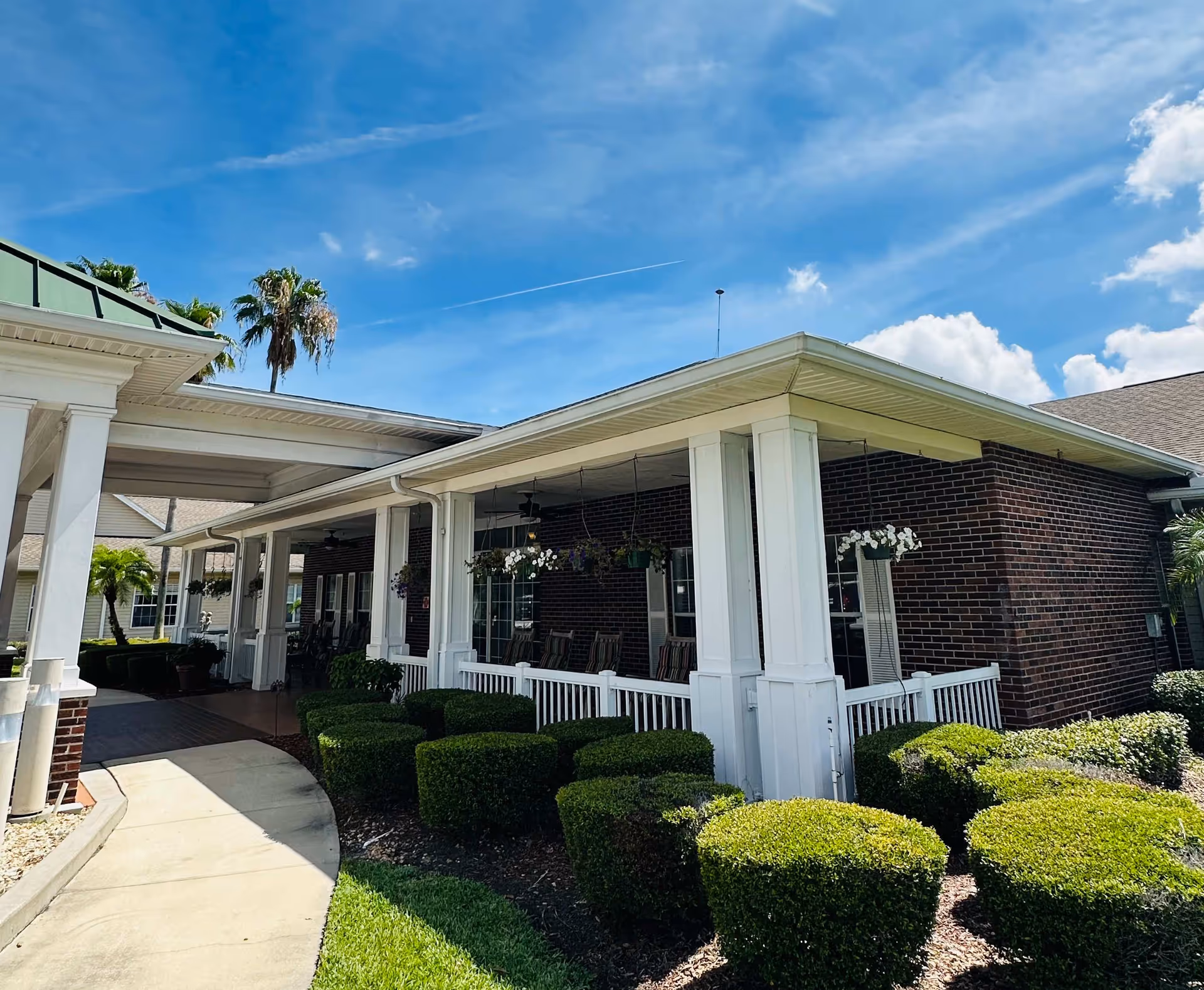 Covered porch and entrance of a brick senior living building with white columns, hanging plants, and manicured shrubs under a blue sky.