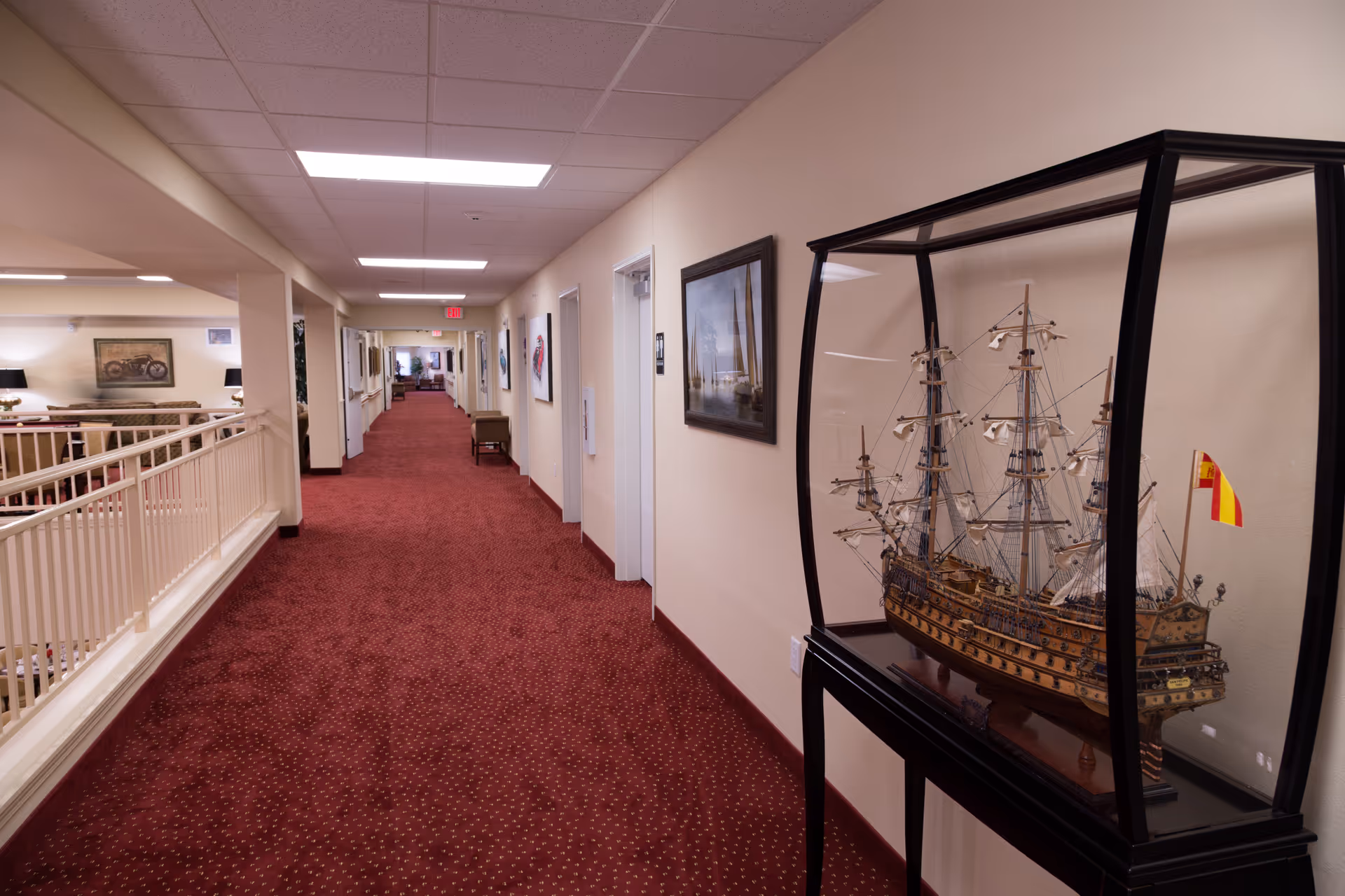 A long indoor hallway with red carpet and cream-colored walls. On the right side, there is a glass display case containing a detailed model ship with sails and rigging. The left side of the hallway has a white railing overlooking a lower level with seating and framed artwork on the walls. The hallway is well-lit with ceiling lights and has several doors and framed pictures along the walls.