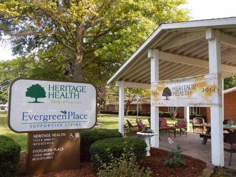 Outdoor view of a supportive living facility named Evergreen Place under Heritage Health. The image shows a large sign with the facility's name and contact information, a covered patio area with chairs and tables, and trees and bushes surrounding the area.