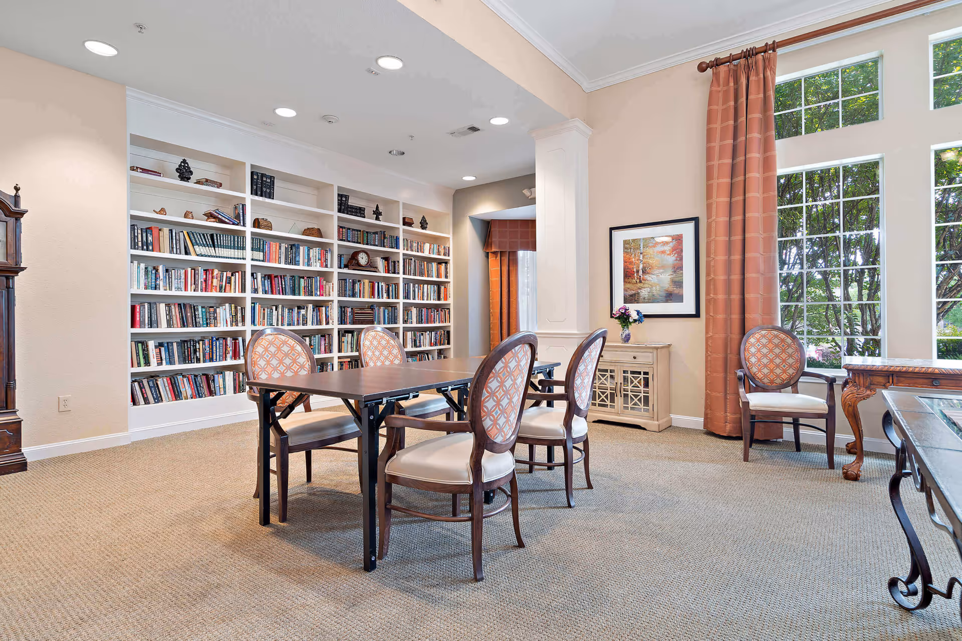 A bright and spacious room with a large bookshelf filled with books along one wall, a table with four upholstered chairs in the center, a window with long curtains letting in natural light, and a framed painting on the wall above a small cabinet with a vase of flowers.