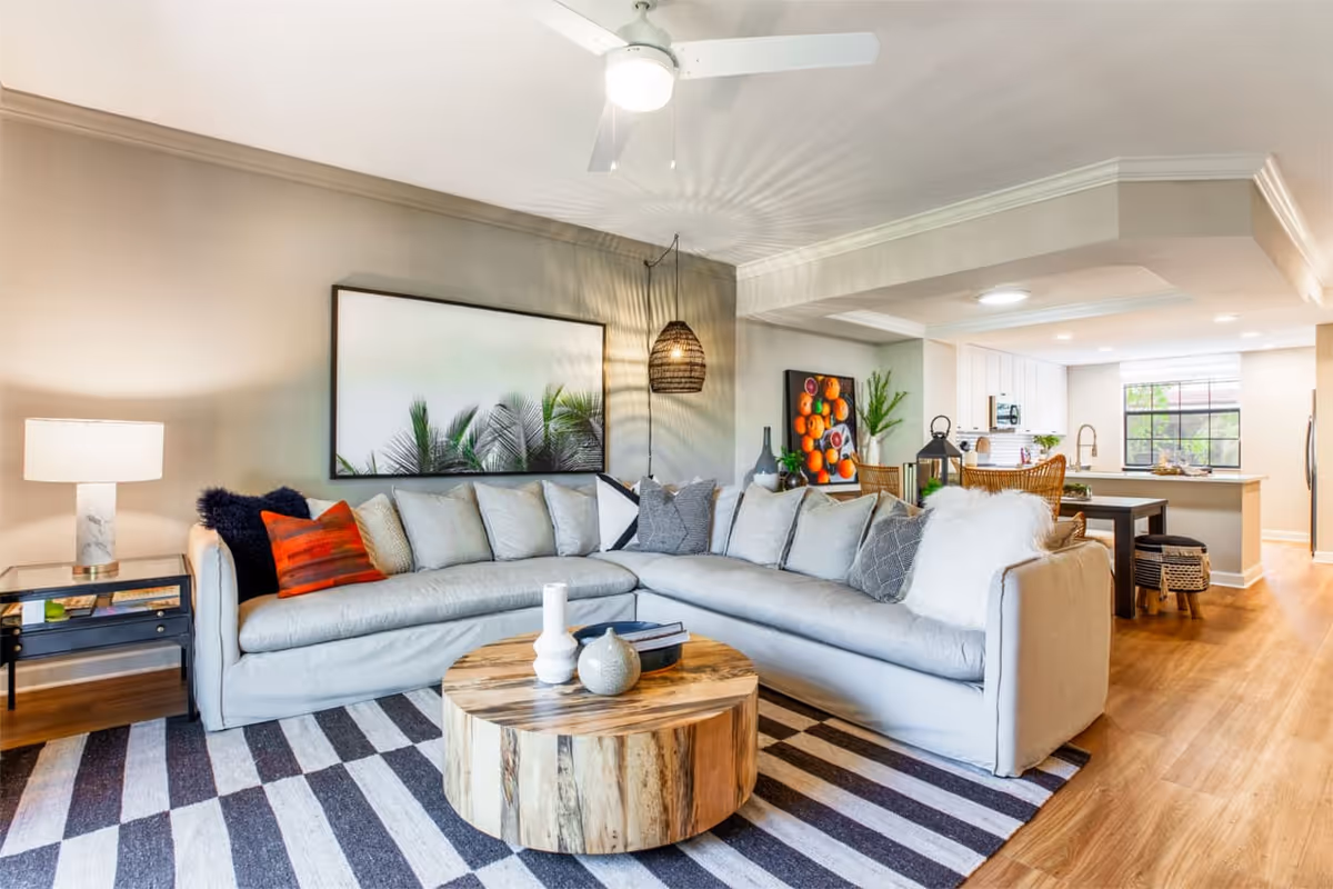 A bright and modern living room featuring a large light gray sectional sofa adorned with various pillows, a round wooden coffee table with decorative items, a striped area rug, a side table with a marble lamp, and wall art depicting green palm leaves. The room opens into a kitchen and dining area with wooden flooring and white cabinetry.
