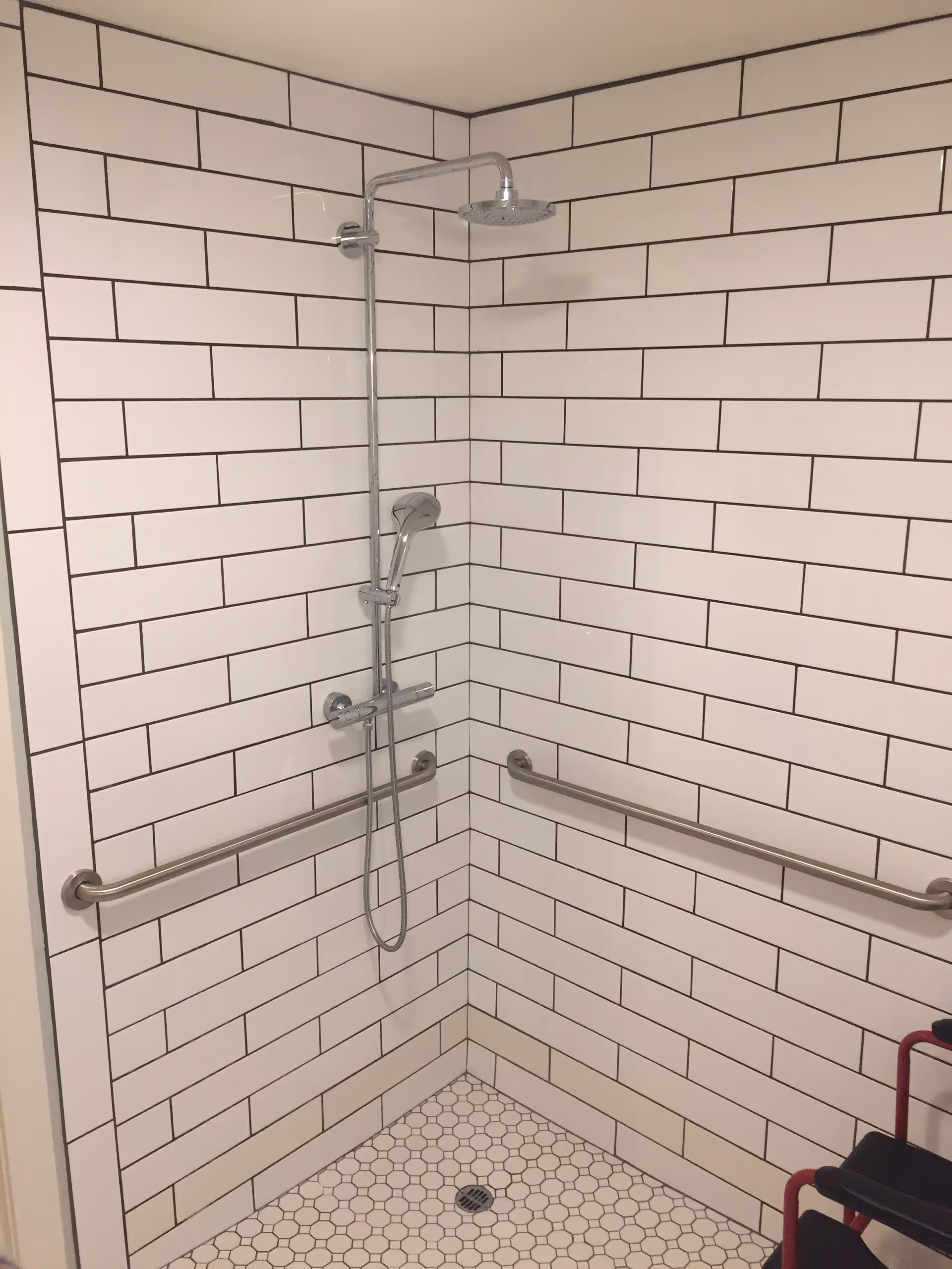 A corner shower area with white subway tiles on the walls and hexagonal white tiles on the floor. The shower has a chrome rain showerhead and a handheld showerhead attached to a vertical bar. There are two horizontal stainless steel grab bars mounted on the walls for support. A black and red shower chair is partially visible in the lower right corner.