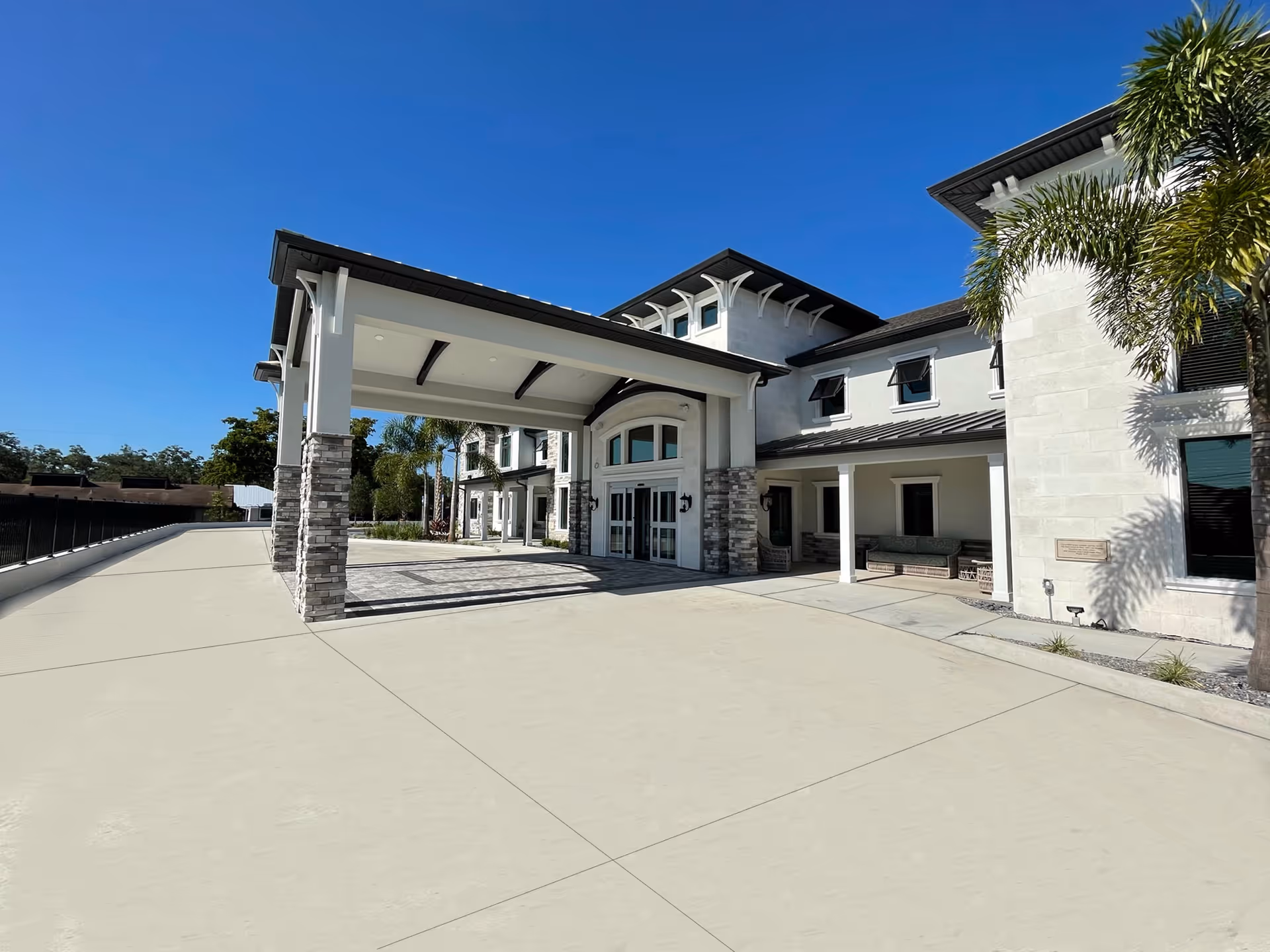 Entrance of a two-story light-colored senior living building with a covered porte-cochere, driveway, and palm trees.