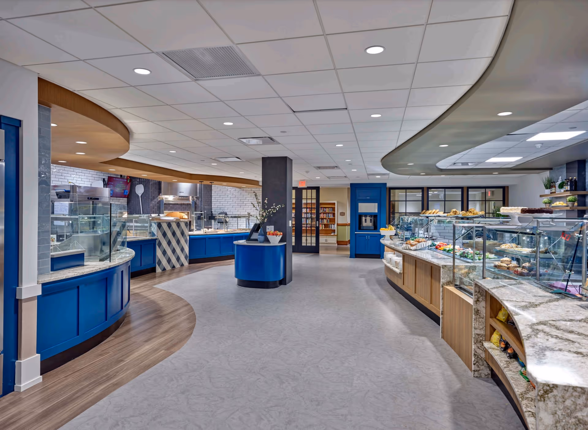 Interior view of a modern dining service area with curved counters and glass food display cases. The counters are blue and wood-toned with marble tops, and the ceiling has recessed lighting. The space is clean and well-lit, with a serving area visible in the background.