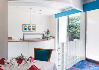 A bright dining area with a table set for a meal, featuring red napkins, glassware, and a floral centerpiece. In the background, there is a counter with a vase of flowers and colorful artwork on the wall. Large windows allow natural light to fill the room and offer a view of greenery outside.