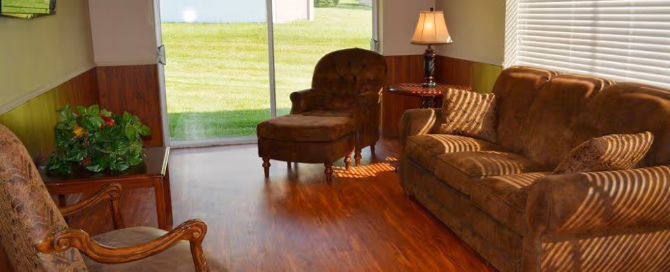 A cozy living room with a brown upholstered sofa and matching armchair with ottoman. There is a wooden side table with a lamp on it next to the sofa, and another wooden chair with patterned upholstery. A sliding glass door opens to a green lawn outside, and sunlight streams through the window blinds, casting shadows on the furniture and wooden floor.