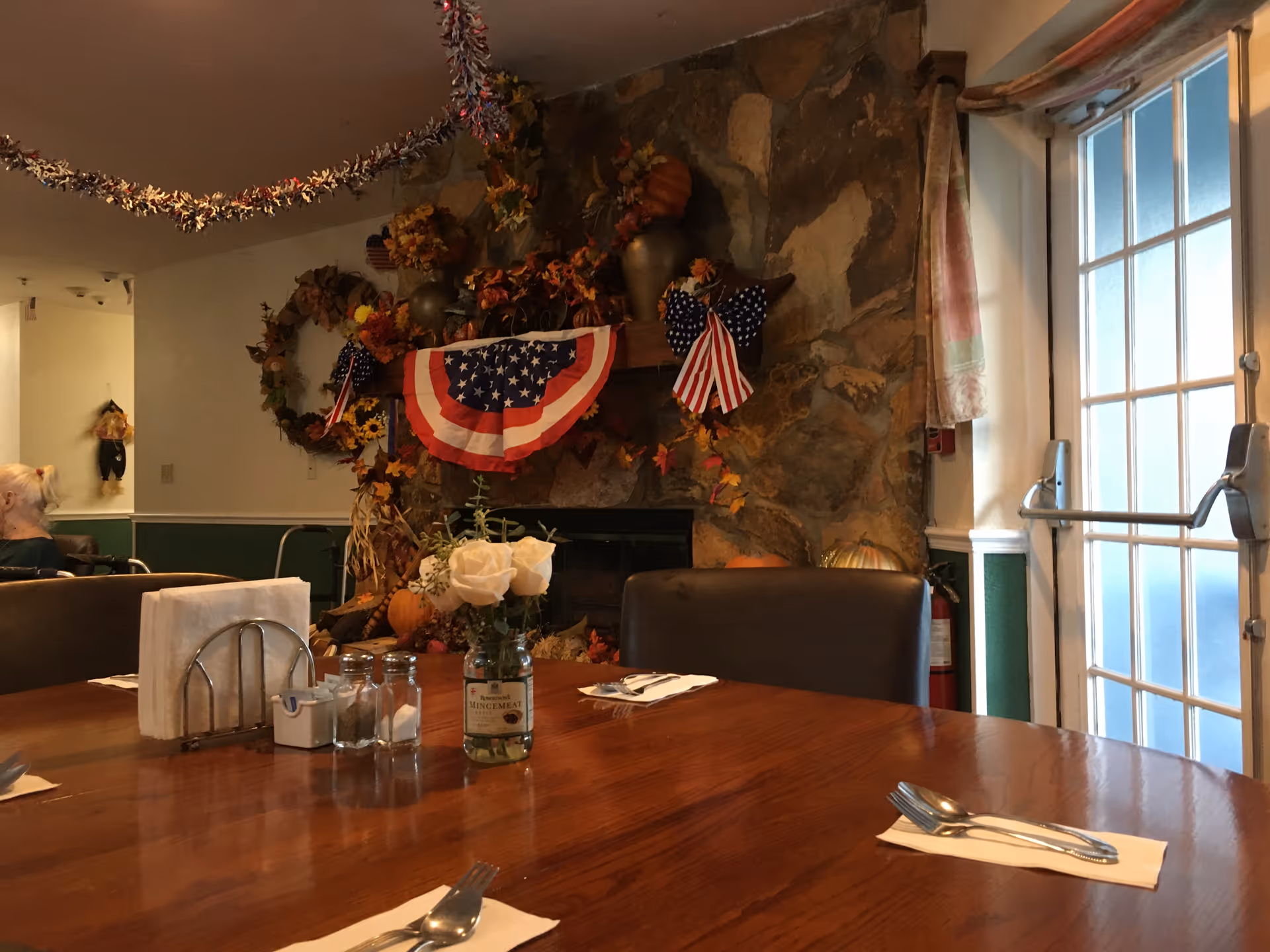 A dining room table set with utensils and napkins in front of a stone fireplace decorated with American flags, fall wreaths, and autumn-themed decorations. A vase with white roses and salt and pepper shakers are on the table. There is a glass door to the right and a person sitting in the background on the left.