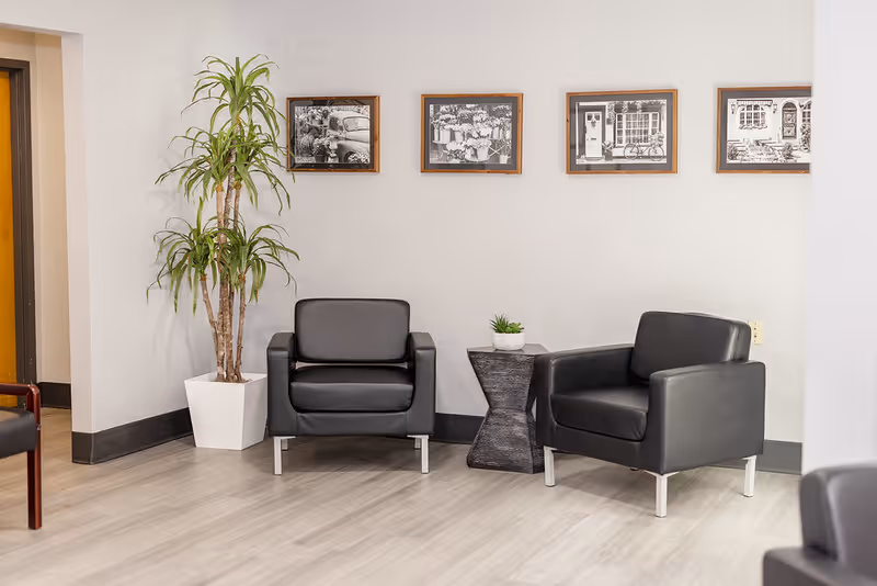 A seating area with two black leather armchairs separated by a small black side table with a small potted plant on top. There is a tall green plant in a white pot to the left, and four framed black and white pictures hanging on the white wall behind the chairs. The floor is light-colored wood.
