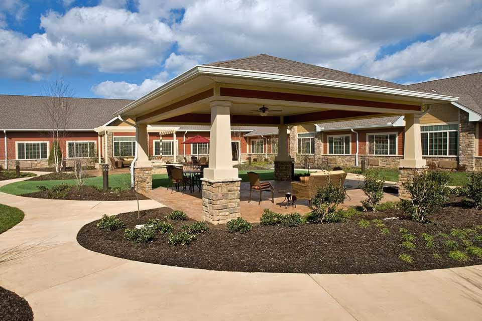 Outdoor covered seating area with stone pillars and a ceiling fan, surrounded by a garden with small bushes and a paved walkway, with a building in the background under a partly cloudy sky.