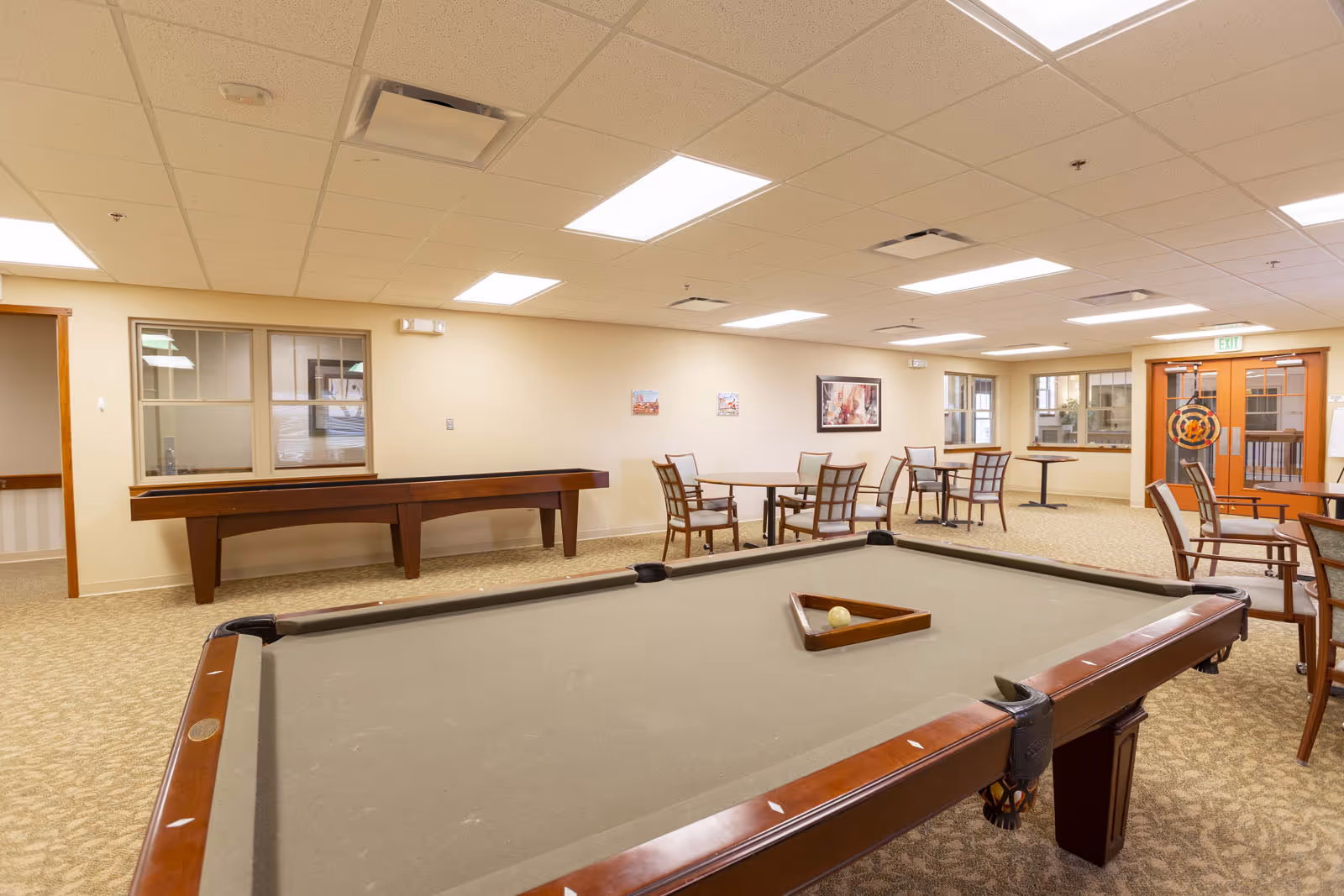 A spacious recreational room with a pool table in the foreground and a shuffleboard table against the wall. Several tables and chairs are arranged around the room, with framed artwork on the walls and large windows letting in natural light. The ceiling has recessed lighting and air vents, and there is a door with glass panels on the right side.