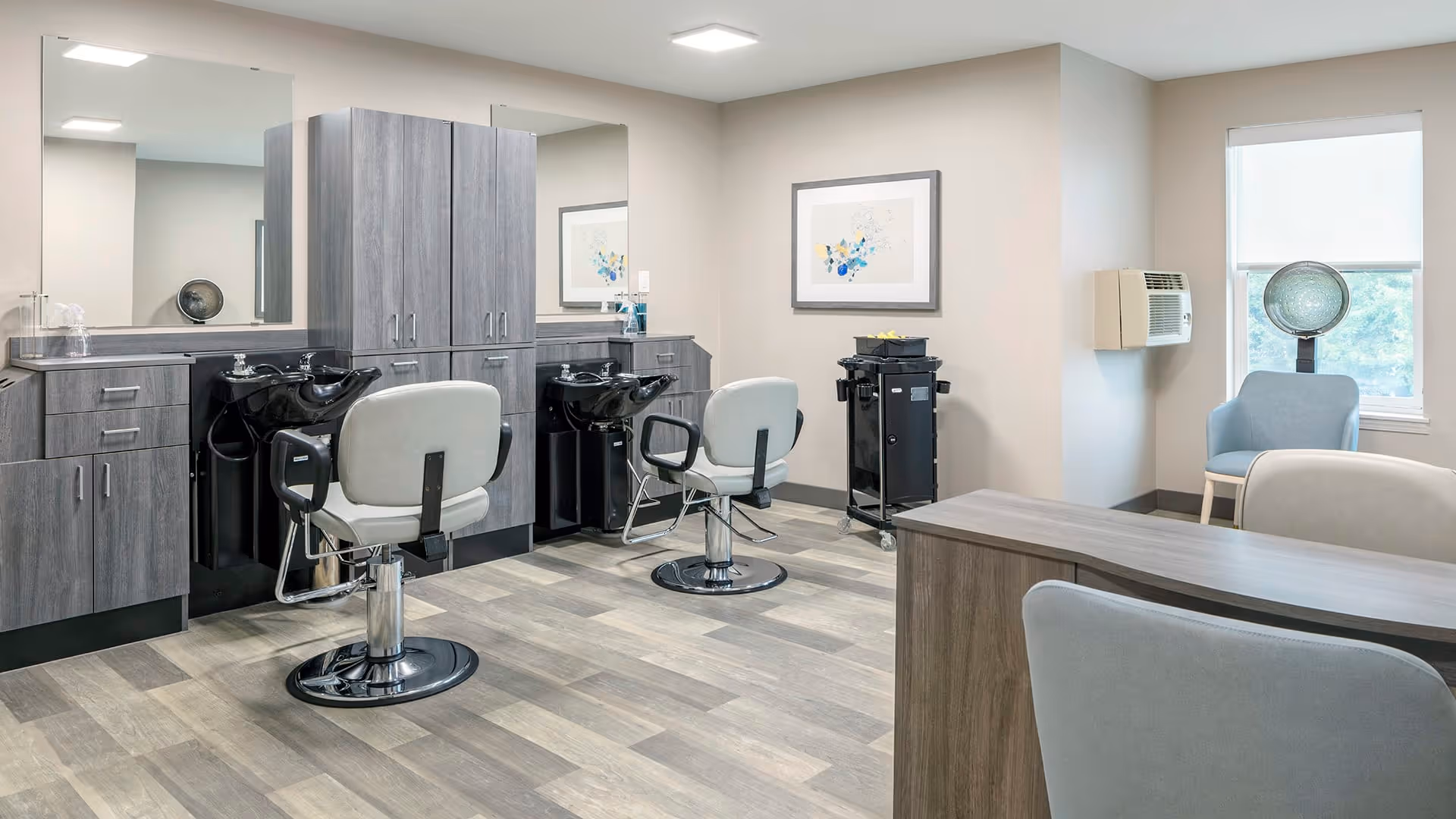 Interior of a salon area in a senior living facility with two salon chairs facing black hair-washing sinks, gray cabinetry with mirrors above, a rolling cart with supplies, a blue chair near a window with a hair dryer hood, and a framed abstract artwork on the wall.