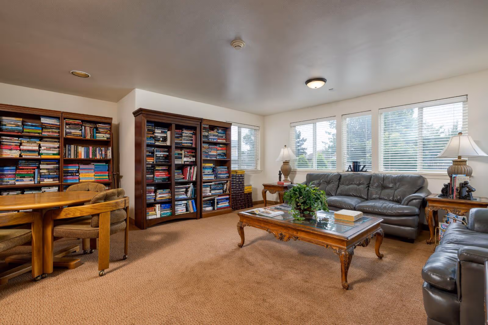 A cozy living room area with two dark leather sofas, a wooden coffee table with a glass top and a potted plant, two wooden bookshelves filled with books, a wooden table with cushioned chairs, and large windows with blinds letting in natural light.