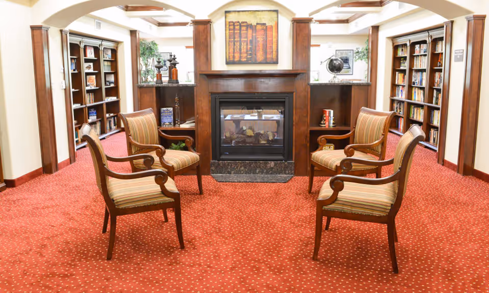 A cozy library or reading room with four wooden armchairs arranged around a central fireplace. The room has red carpet with a dotted pattern, wooden bookshelves filled with books on either side, and a painting of books above the fireplace. There are decorative items on the shelves and a globe on the right side.