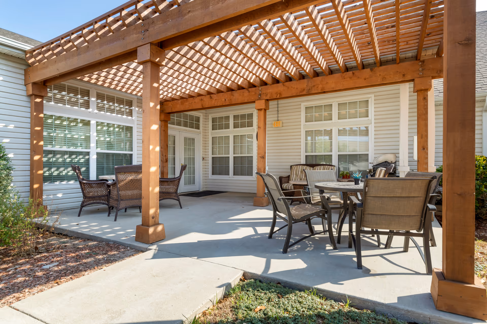 Covered outdoor patio with a wooden pergola, tables and chairs next to a light-colored building.