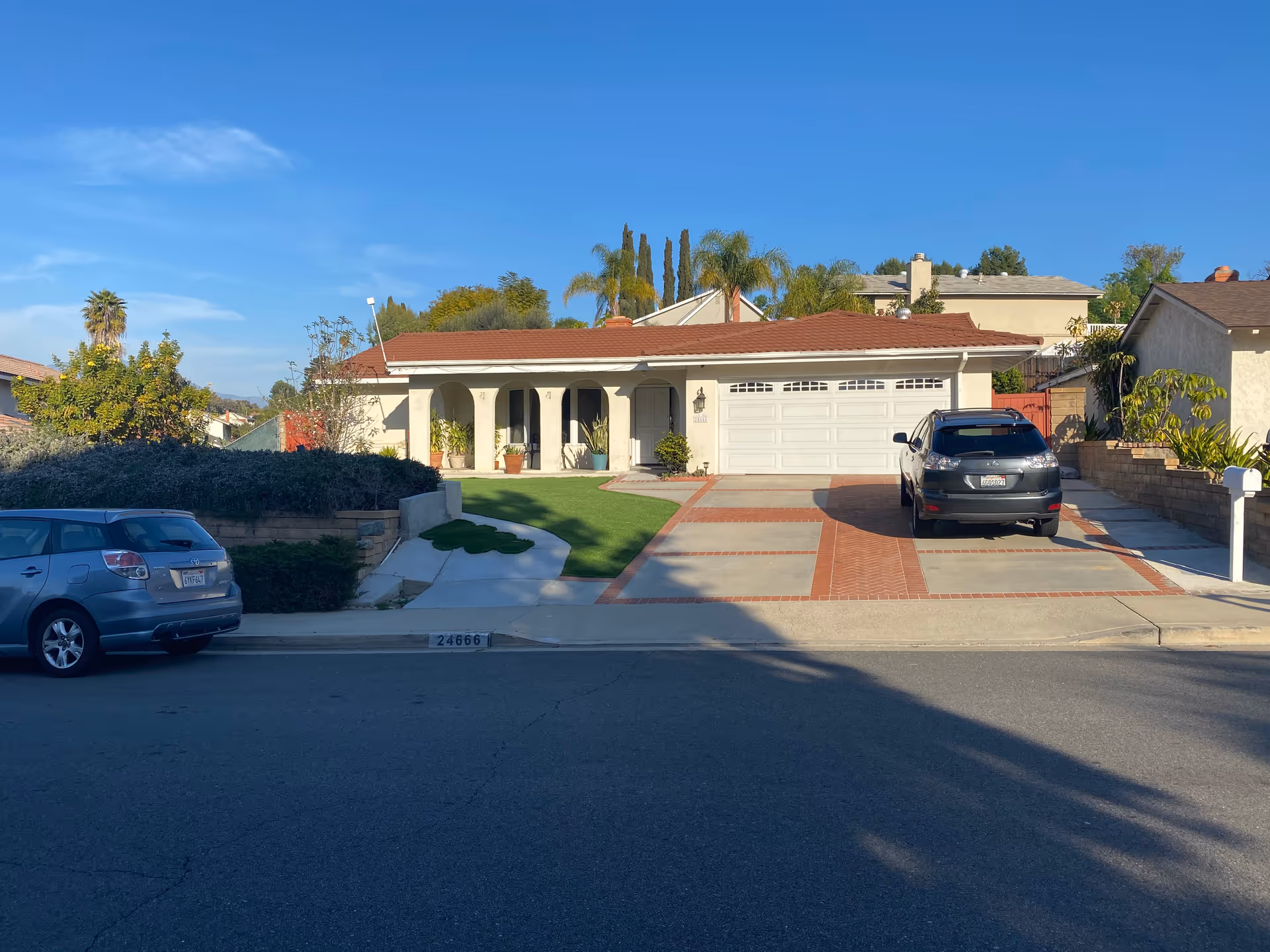 Front exterior view of a single-story house with a red-tiled roof, white garage door, and a driveway with a parked black SUV. The house has a small front porch with potted plants and a well-maintained lawn. There are trees and shrubs around the property, and a clear blue sky overhead.
