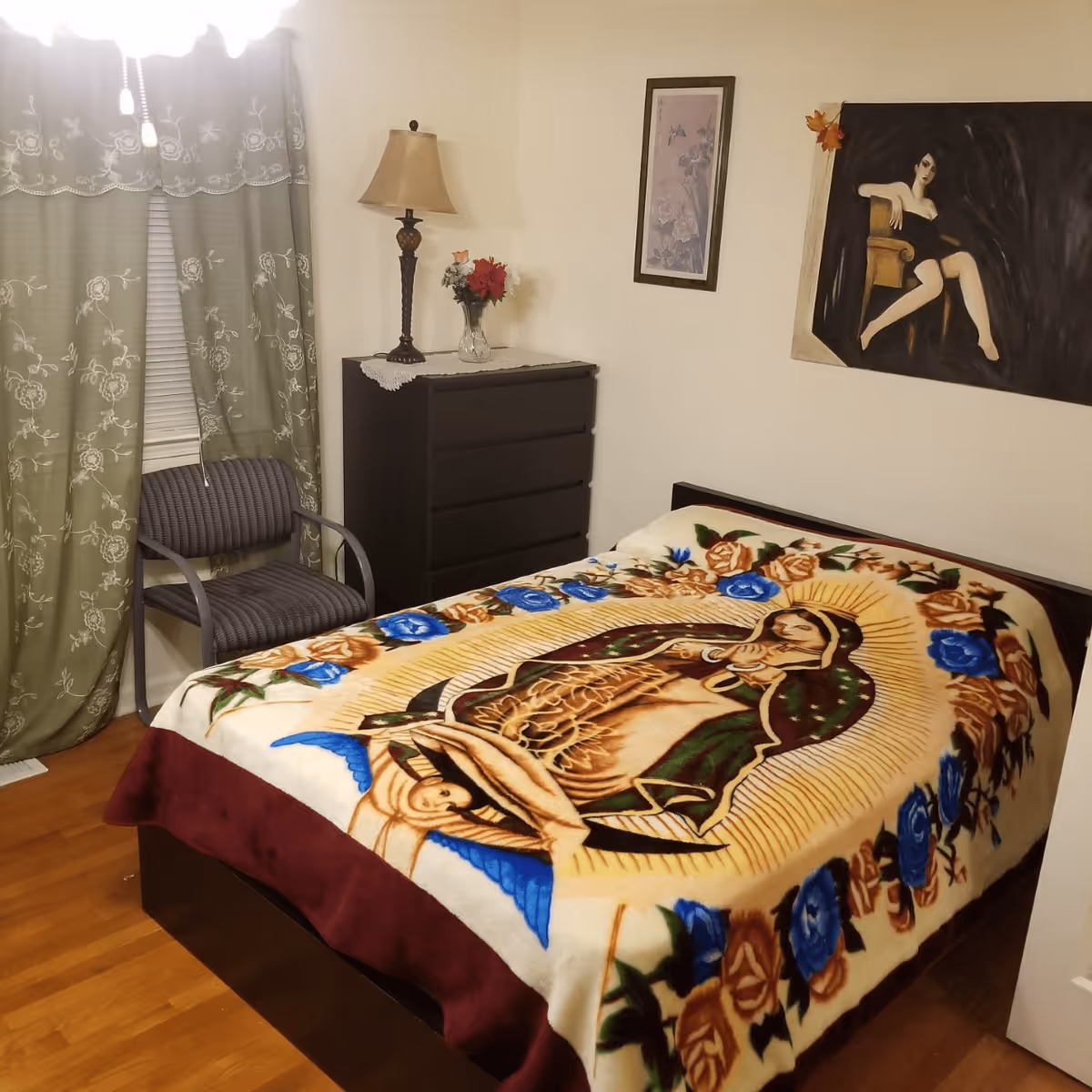 A bedroom with a bed covered by a colorful blanket featuring a religious image, a black dresser with a lamp and a vase of flowers on top, a gray chair near a window with green floral curtains, and two framed artworks on the walls.